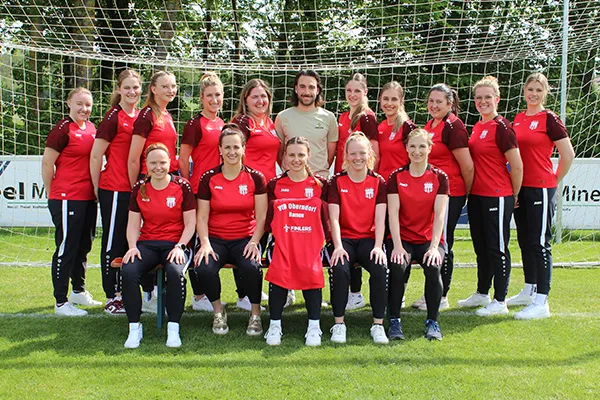 Das Bild zeigt die Damenmannschaft des VFB Oberndorf, die zusammen vor einem Fußballtor auf einem Rasenfeld stehen und sitzen. Die Teammitglieder tragen rote Shirts mit dem Logo des VFB Oberndorf und schwarze Hosen, und sie lächeln in die Kamera. Eine Person in der Mitte hält ein rotes Shirt mit dem Logo des VFB Oberndorf und dem Namen des Sponsors "FinLers GmbH".