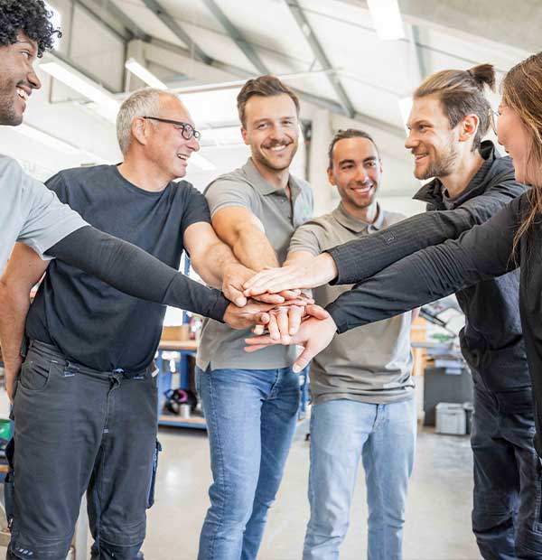 6 Personen lächeln und berühren in der Mitte ihre Hände und zeigen so ihren Zusammenhalt.