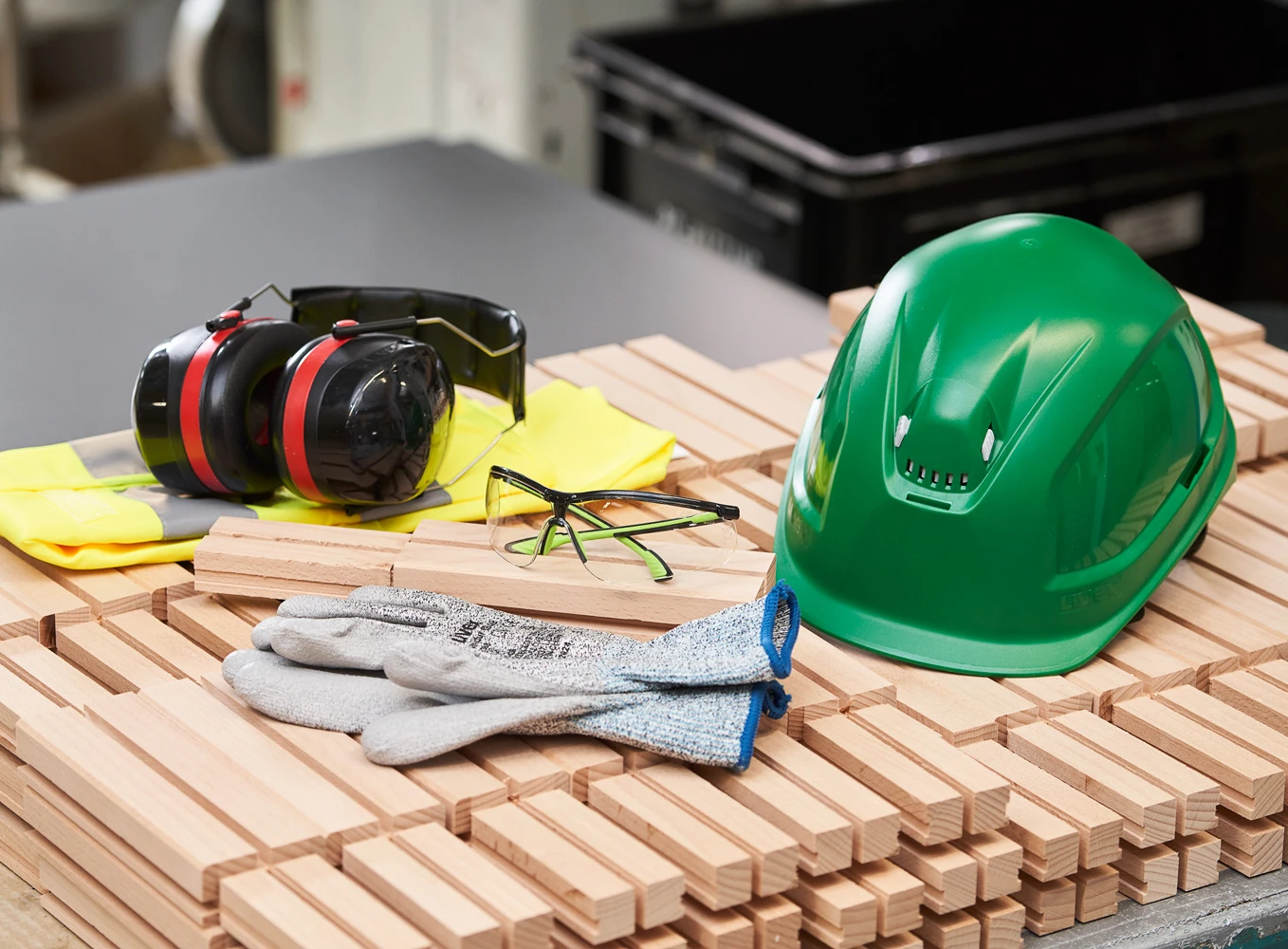 Schutzausrichtung bestehend aus Helm, Ohrschutz, Handschuhen und einer Schutzbrille liegt auf Holzteilen in einer Werkstatt.