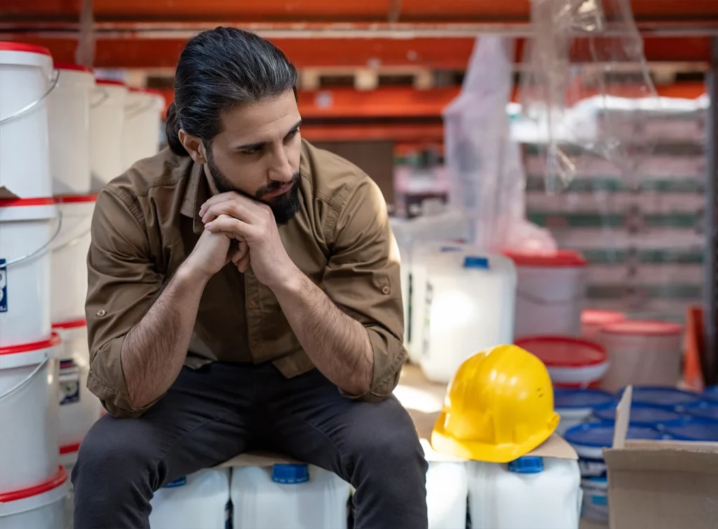 Ein nachdenklicher Mann in Arbeitskleidung sitzt in einem Lager zwischen Farbeimern und einem gelben Schutzhelm.