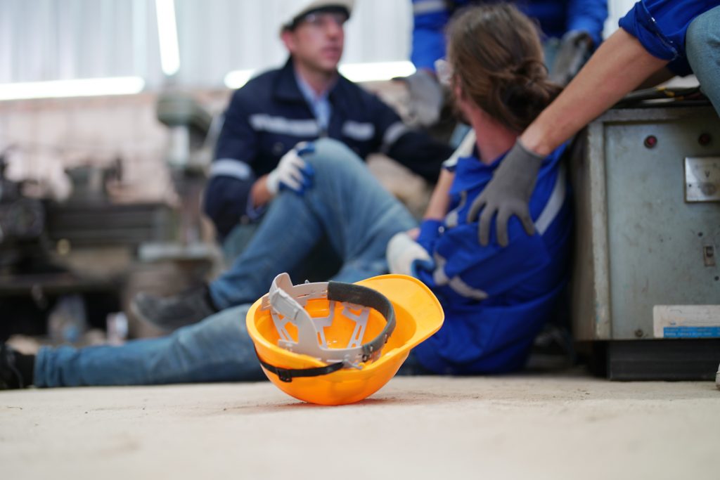 Arbeitnehmer hat einen Arbeitsunfall und liegt verletzt auf dem Boden. Seine Arbeitskolleg:innen helfen ihm.